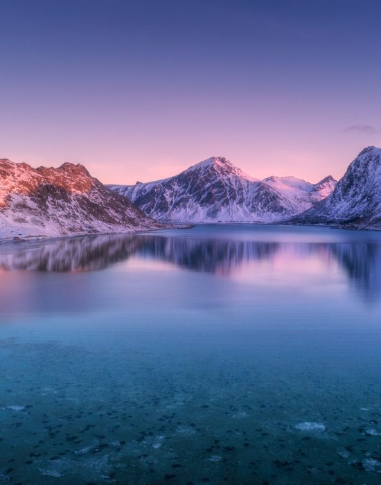 Aerial view of snow covered mountains and colorful sky reflected in water at dusk. Winter landscape with sea, snowy rocks, purple sky, reflection at sunset. Lofoten islands, Norway at twilight. Nature